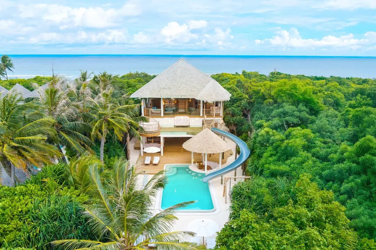 A luxury Water Retreat with Slide, this villa features a thatched roof, private pool, and waterslide, nestled among lush green trees with the ocean visible in the background beneath a blue sky with scattered clouds.