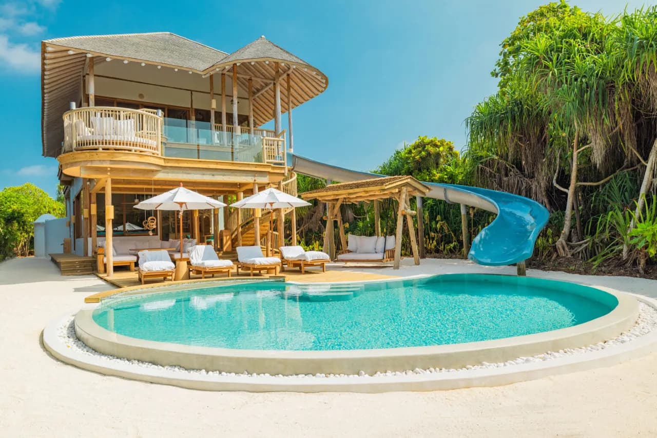 A luxurious beachfront villa with a round pool, wooden lounge chairs under white umbrellas, a water slide, and lush tropical greenery under a clear blue sky.