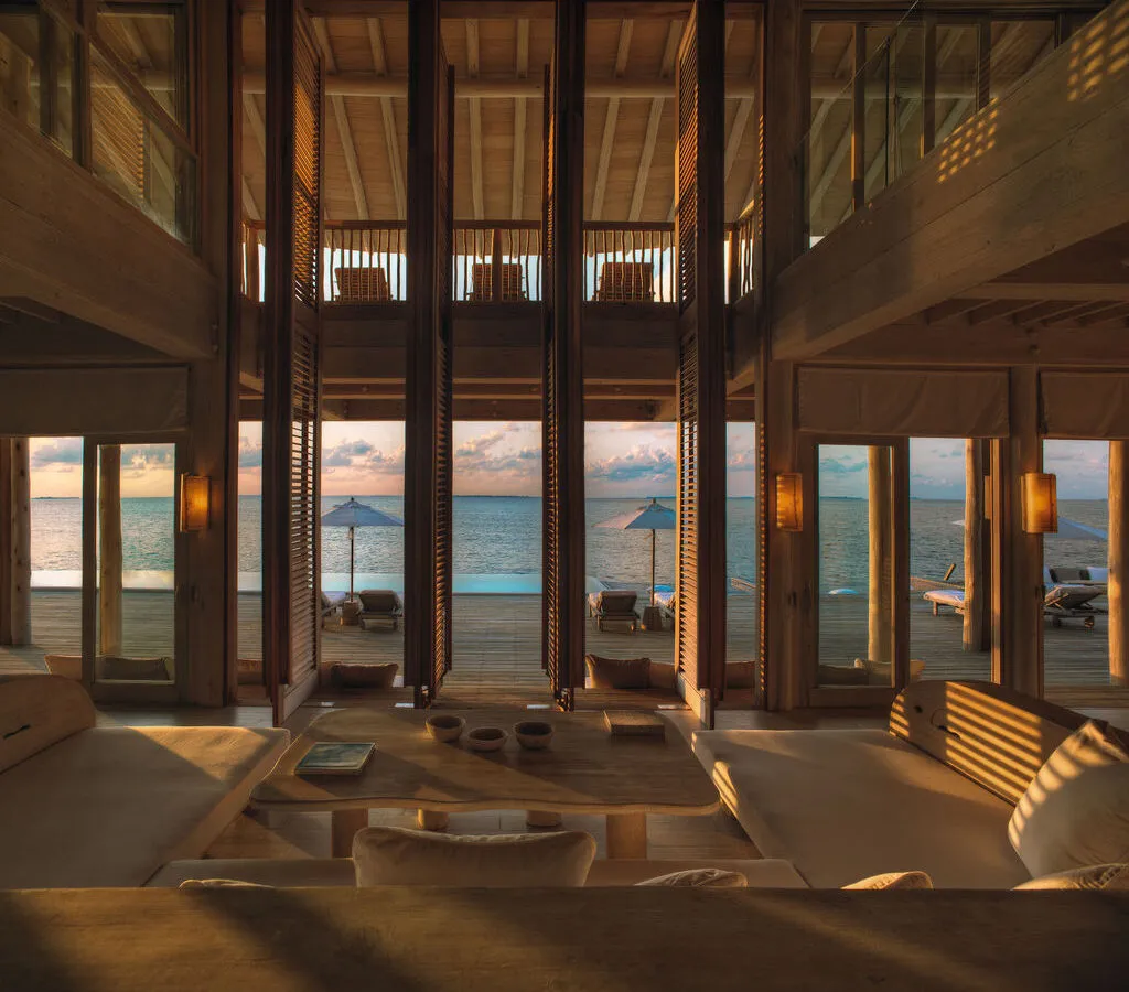 Luxurious open-plan beachfront villa with wooden interiors, large windows, and sliding doors opening to a deck with sun loungers, parasols, and a view of the sea at sunset. Warm sunlight fills the space.
